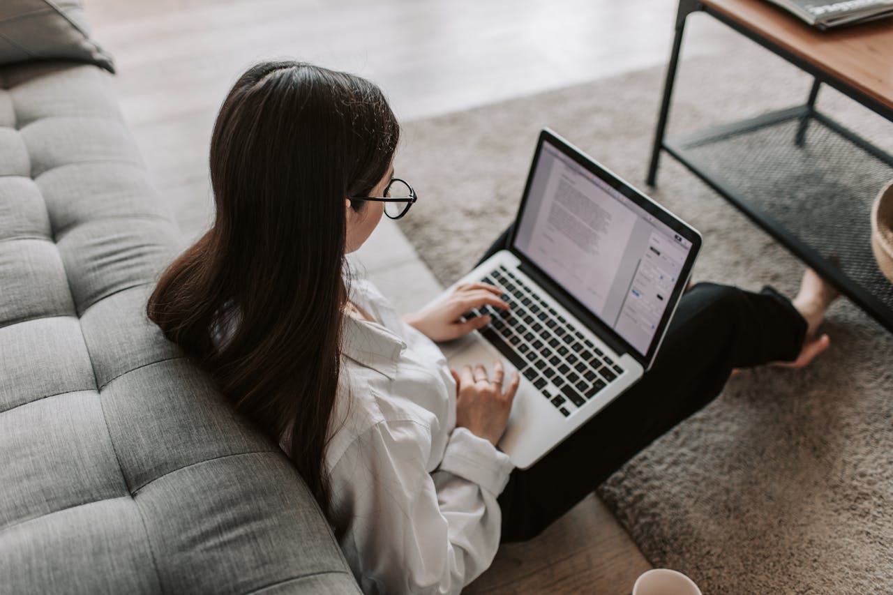 gallery-05 Woman working remotely with a laptop on the floor next to a sofa, enjoying comfortable home office setup.