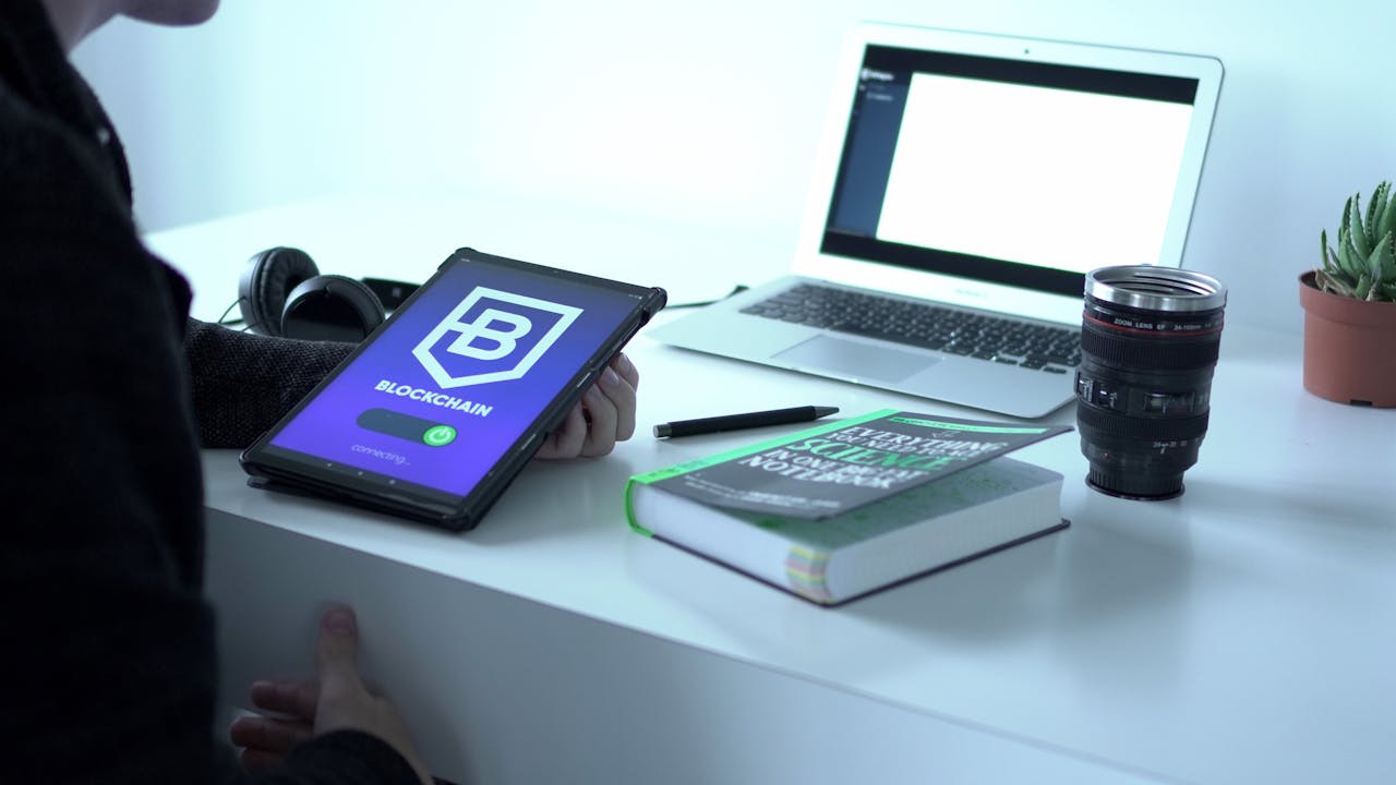 our-story A desk with tablet displaying blockchain, laptop, book, and camera lens, symbolizing tech and innovation.
